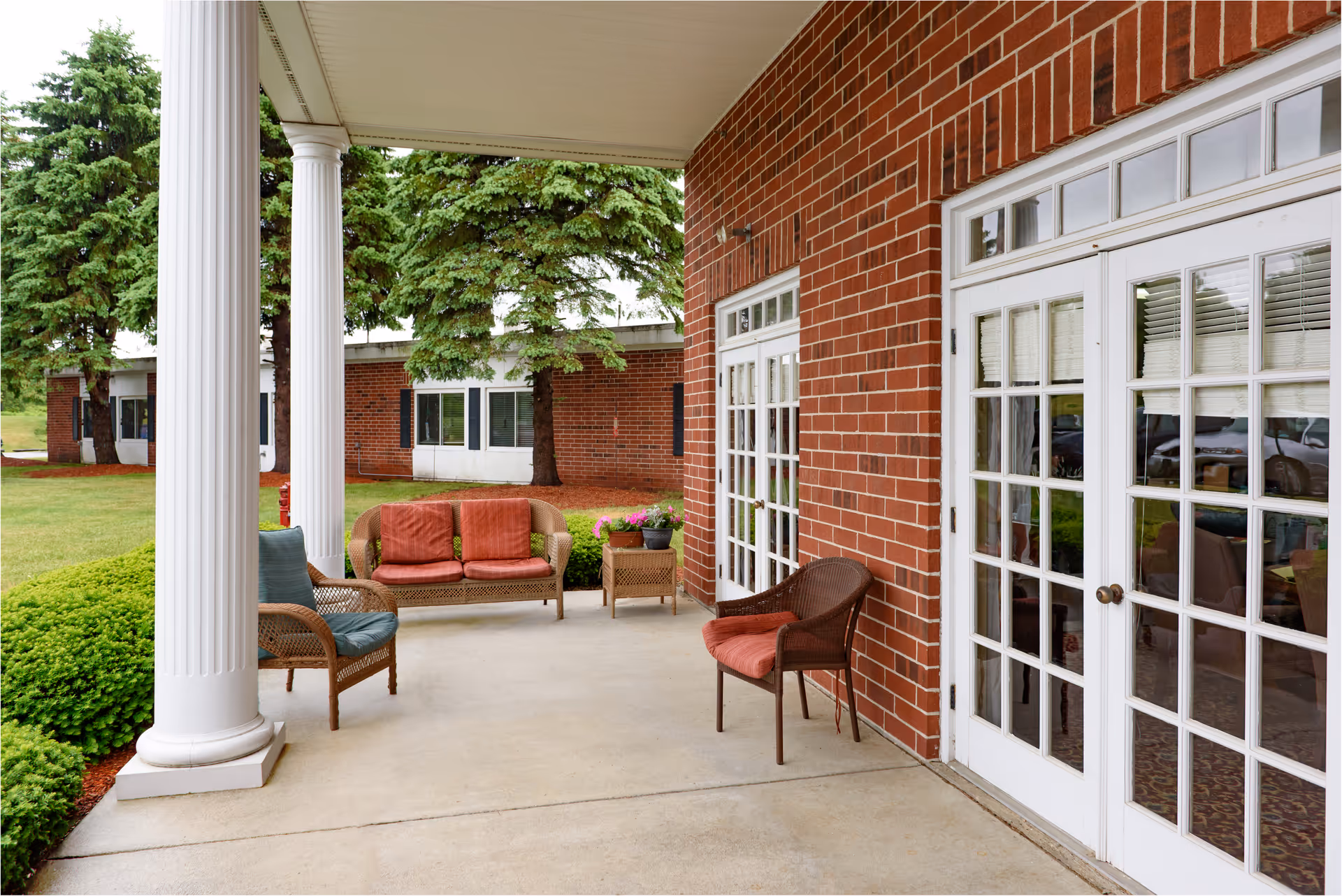 Covered outdoor patio area with white columns, wicker chairs with cushions, a small table with potted plants, and a brick wall with white-framed glass doors leading inside. Green bushes and trees are visible in the background.