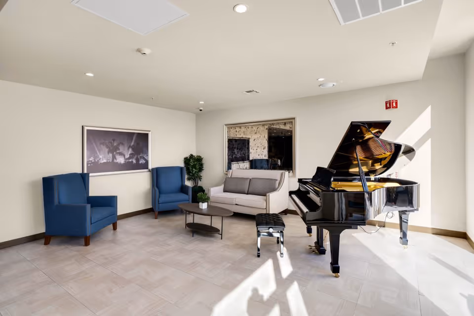 Sunlit senior living lounge with a grand piano, a sofa, two blue armchairs, and a coffee table.