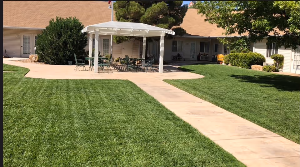 Outdoor courtyard area with a concrete walkway leading to a covered patio with tables and chairs. Surrounding the patio is well-maintained green grass, shrubs, and trees. The background shows the exterior of a single-story building with multiple doors and windows.