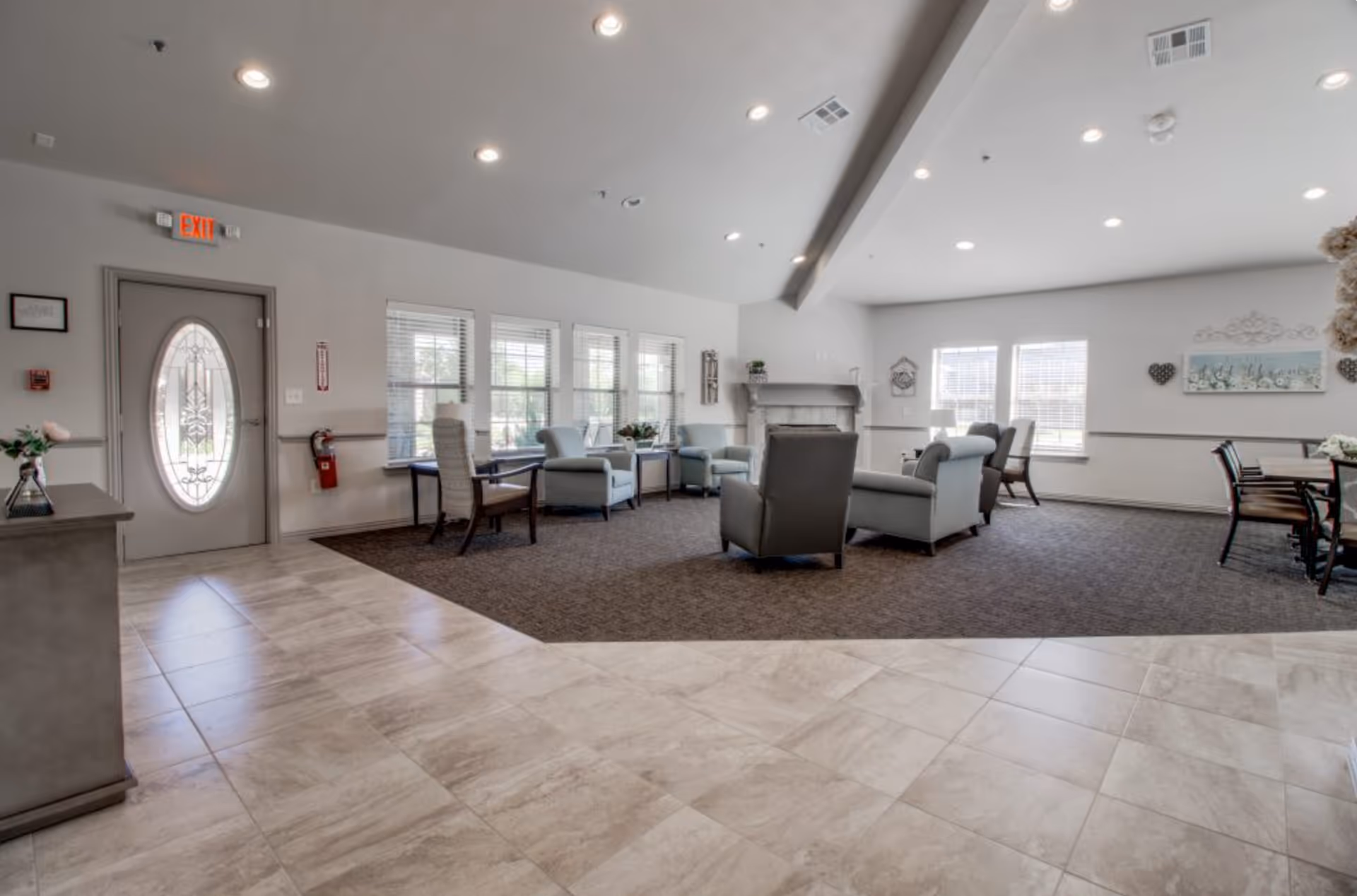A spacious and well-lit common living area in an assisted living facility featuring multiple armchairs arranged around a fireplace, large windows with blinds, a tiled floor transitioning to carpet, and a dining table with chairs in the background.