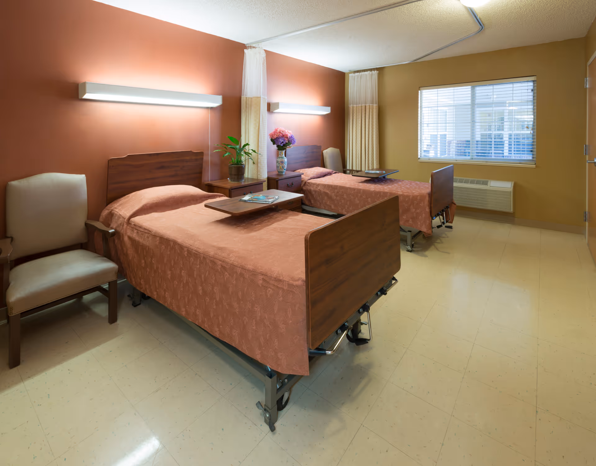 A room with two hospital-style beds covered in matching brown bedspreads, each with an overbed table. There is a beige armchair to the left, a nightstand with a plant and a vase of flowers between the beds, and a window with blinds on the right wall. The walls are painted in warm tones of brown and beige, and there are wall-mounted lights above each bed.