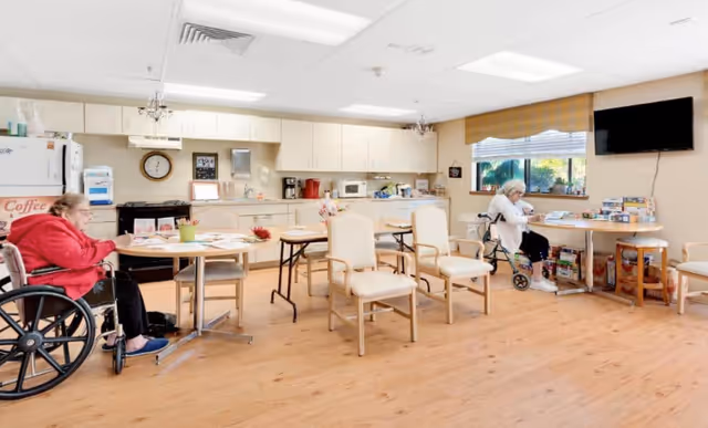 A bright common area in a senior living facility with light wood flooring and cream-colored walls. Two elderly women are seated, one in a wheelchair at a round table on the left and another on a walker near a window on the right. The room features a kitchenette with white cabinets, a refrigerator, microwave, coffee maker, and various kitchen items. Several chairs and tables are arranged around the room, and a flat-screen TV is mounted on the wall near the window.
