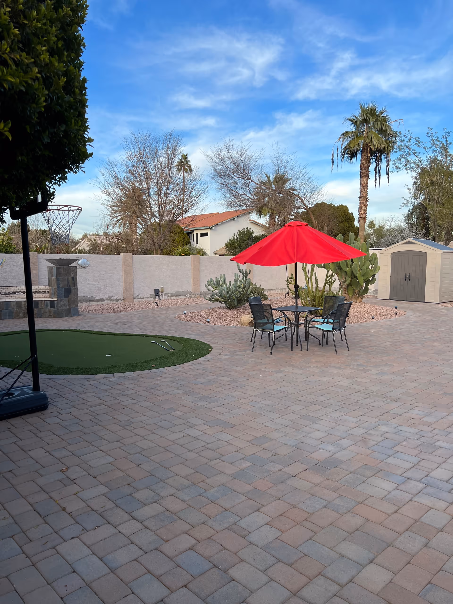 Paved backyard patio with a round table and red umbrella, desert landscaping, palm trees and a storage shed.