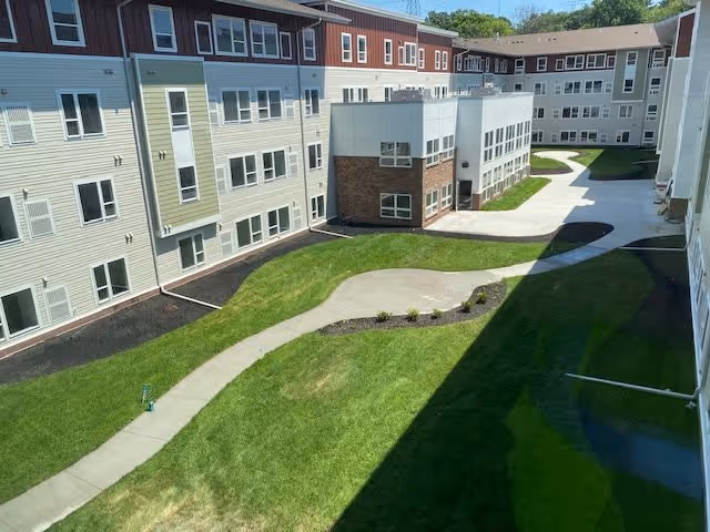 View of a senior living facility courtyard with a winding concrete pathway surrounded by green grass and small plants, bordered by a three-story building with multiple windows and a mix of beige, white, and brick exterior walls under a clear blue sky.