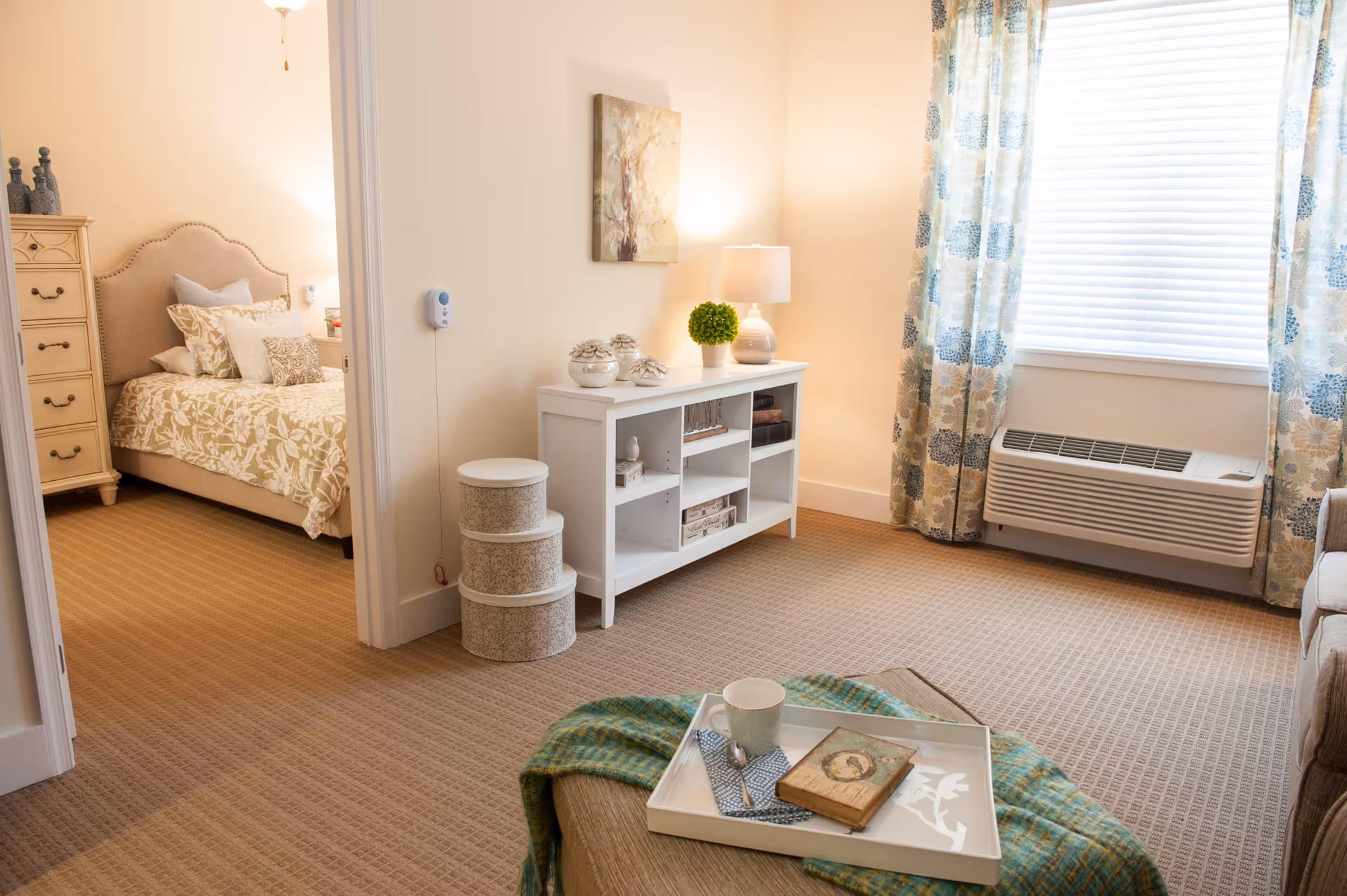 Sunlit living room featuring a tray on an ottoman, white console shelving, patterned curtains, an air conditioner under the window, and an adjacent bedroom visible through a doorway.