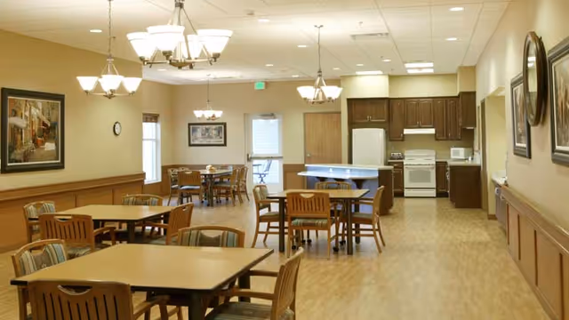 A spacious dining area in a senior living facility with several wooden tables and chairs arranged neatly. The room features warm lighting from ceiling chandeliers, framed artwork on the walls, and a kitchen area in the background equipped with a refrigerator, stove, microwave, and cabinets. The floor is covered with light wood-style flooring, and there are windows and a door at the far end of the room.