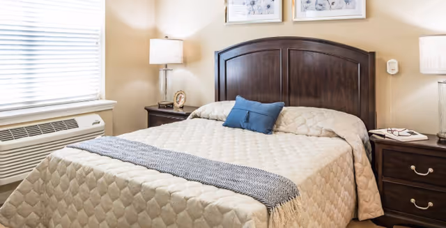 A neatly made bedroom with a dark wooden headboard, bedside tables and lamps, a window with blinds and an air-conditioning unit.