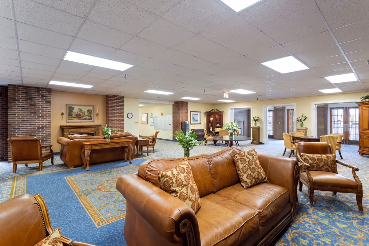 A spacious senior living facility common area with brown leather sofas and armchairs, patterned blue and gold carpet, wooden tables with floral centerpieces, brick pillars, and framed artwork on the walls under bright ceiling lights.