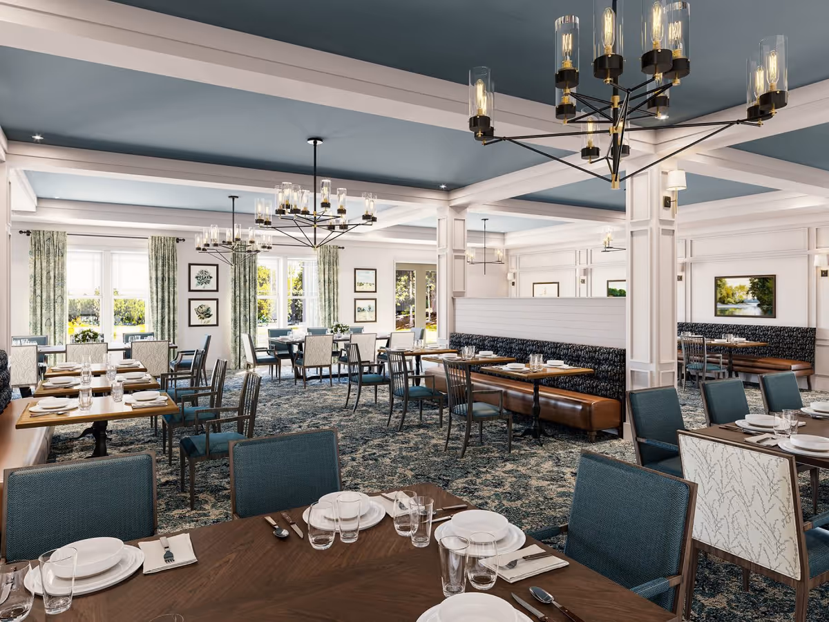 A spacious and elegant dining room with multiple tables set with white plates, glasses, and silverware. The room features blue and white upholstered chairs, patterned carpet, large windows with green floral curtains, and modern chandeliers hanging from a blue ceiling. Artwork and framed pictures adorn the walls.