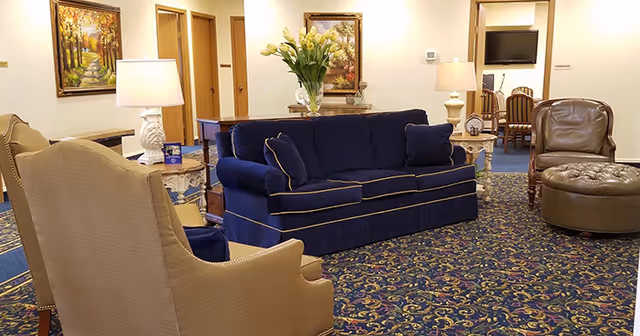 A cozy lobby seating area featuring a navy blue sofa with decorative pillows, two armchairs, and a coffee table. The room is adorned with floral arrangements and artwork on the walls, creating a welcoming atmosphere.
