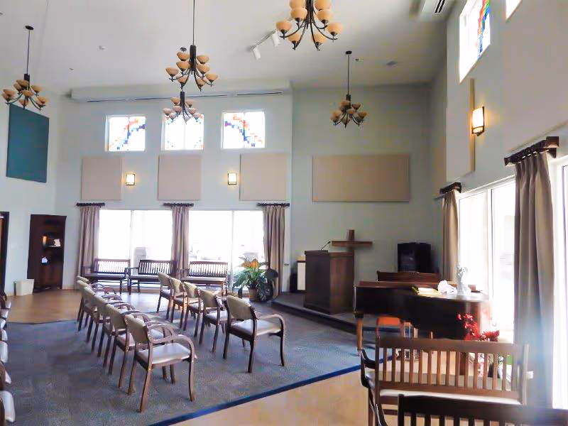 A bright chapel-style common room with rows of chairs facing a podium and piano under chandeliers and stained-glass windows.