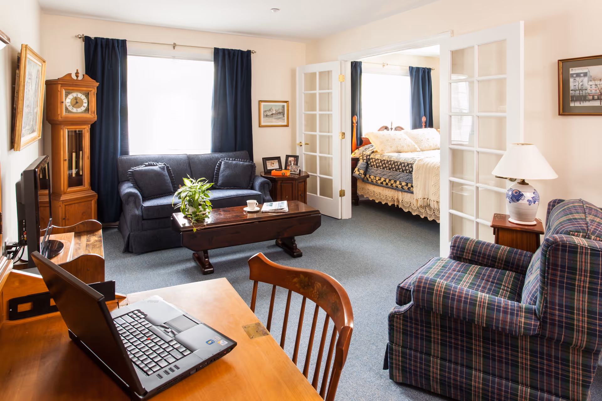 Cozy living room with a sofa, coffee table, armchair and a desk with a laptop, opening to a bedroom through French doors.