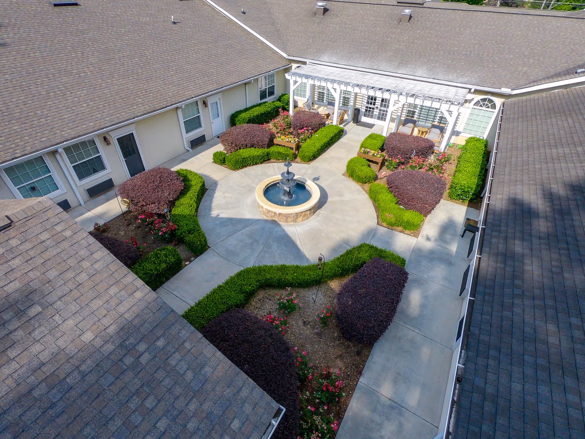 Aerial view of a landscaped courtyard with a central round fountain, concrete walkways, trimmed shrubs, and surrounding building with a pergola and seating.