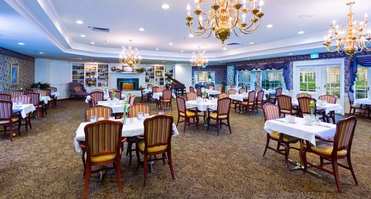 A spacious dining room in an assisted living community with multiple tables covered in white tablecloths, each surrounded by wooden chairs with striped upholstery. The room features ornate chandeliers, a patterned carpet, a fireplace with built-in bookshelves, a grand piano, and large windows with purple drapes allowing natural light to enter.