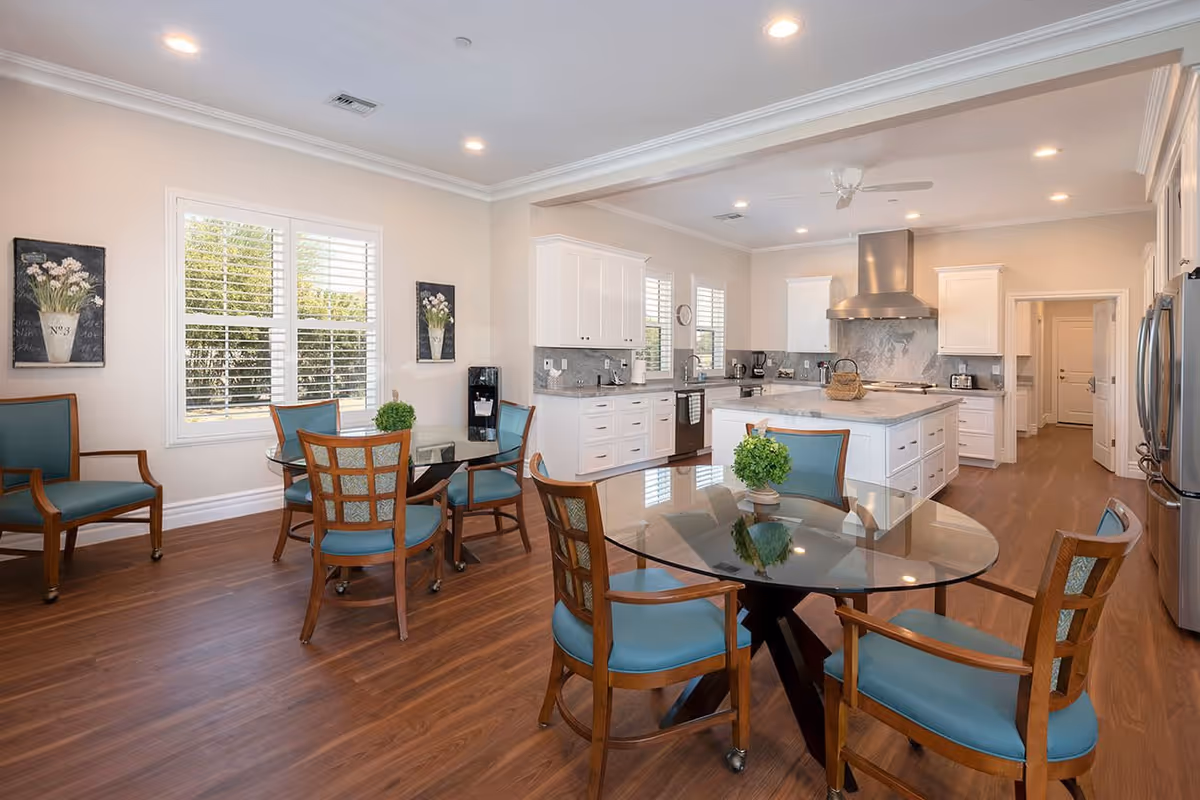 A bright and spacious kitchen and dining area with wooden floors, white cabinetry, and stainless steel appliances. There are two glass-top dining tables with wooden chairs featuring blue cushions. The room has large windows with white shutters, recessed lighting, and decorative wall art with floral designs.