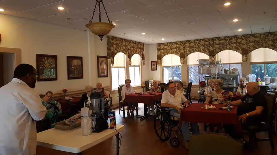 A group of elderly residents seated at tables with red tablecloths in a dining room of an assisted living community. A staff member in a white coat stands near a table with a blender and other items, engaging with the residents. The room has large windows with floral valances, framed artwork on the walls, and a wooden floor.