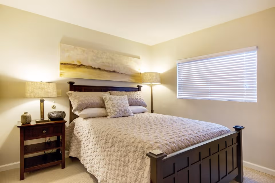 A cozy bedroom with a large wooden bed frame, neatly made with a beige quilt and multiple pillows. There is a wooden nightstand on the left side with a lamp and decorative items. A floor lamp stands in the corner near a window with closed blinds. A landscape painting hangs above the bed on a light-colored wall.