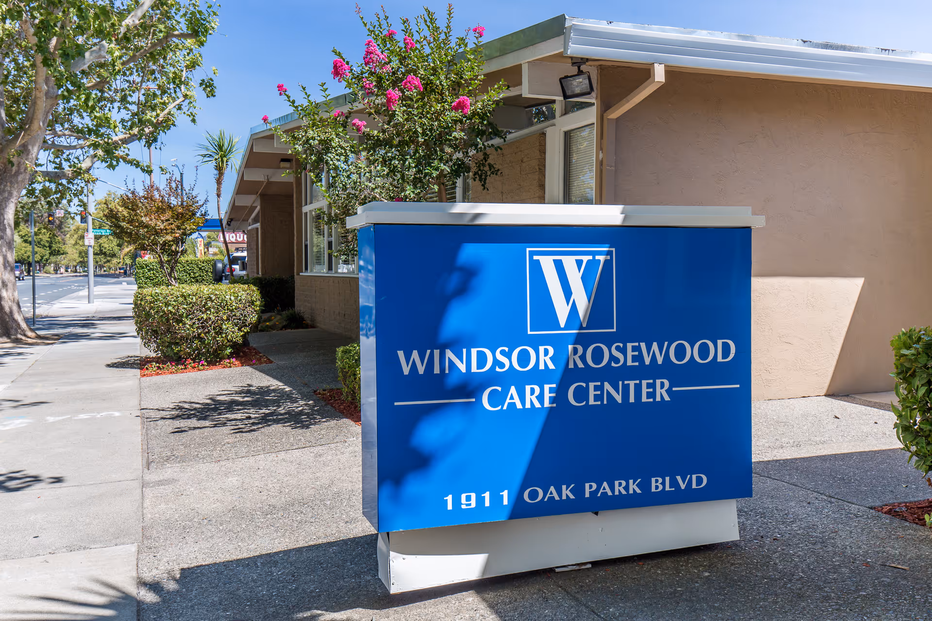 Outdoor view of the Windsor Rosewood Care Center sign in front of the building, with trees, bushes, and a sidewalk along the street.