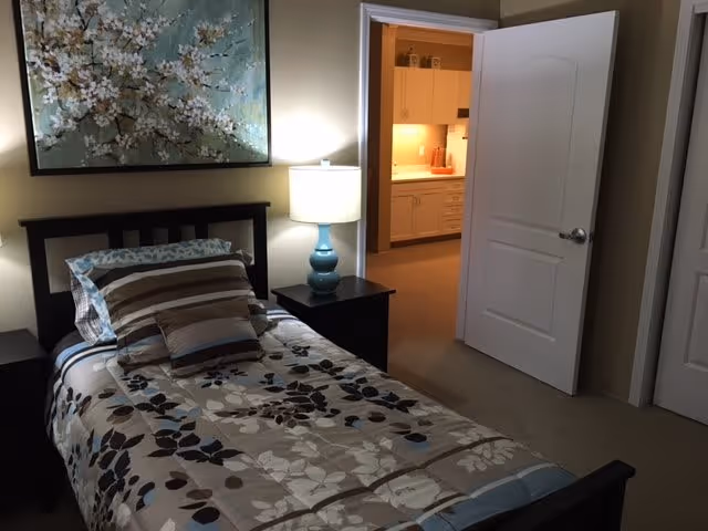 A neatly made single bed with floral bedding and striped pillows in a small bedroom with a nightstand, lamp, and an open door revealing a kitchenette.