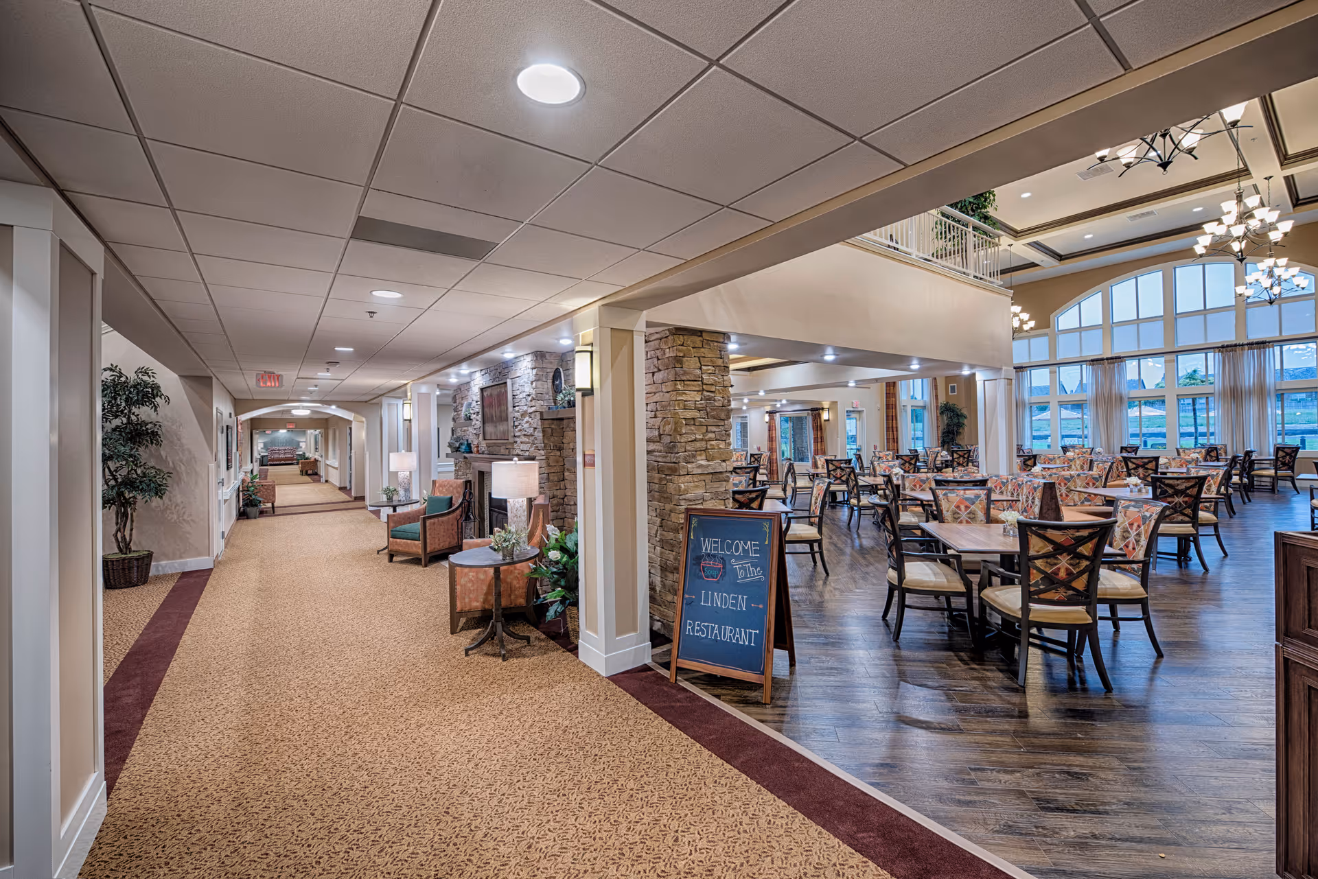 Spacious interior corridor opening into an airy dining room with tables and chairs in a senior living facility.