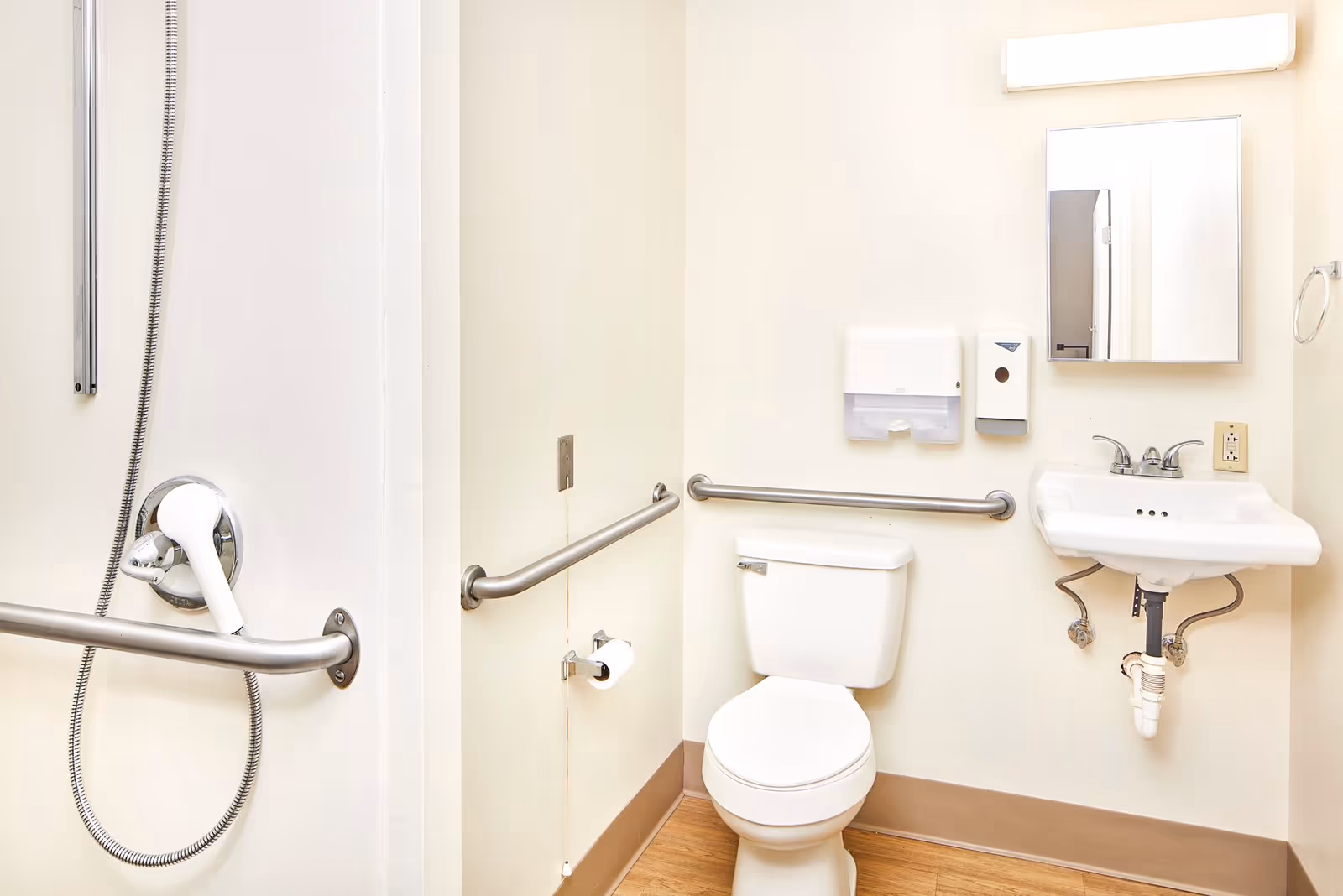 Accessible bathroom with a toilet, wall-mounted sink, mirror, grab bars and a handheld shower head.
