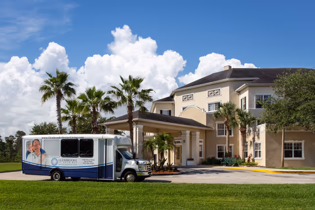 Exterior view of Glenbrooke Senior Living facility with a beige multi-story building, palm trees, and a shuttle bus parked in front under a covered entrance. The sky is blue with scattered white clouds.
