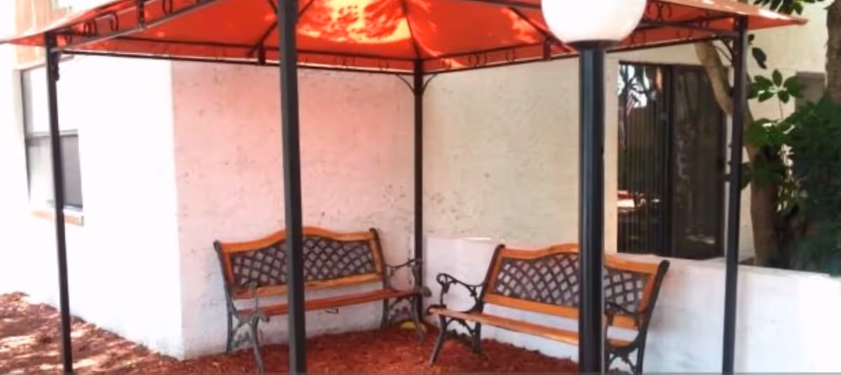 Two wooden-and-metal benches sit under a red canopy beside a white building wall and windows.