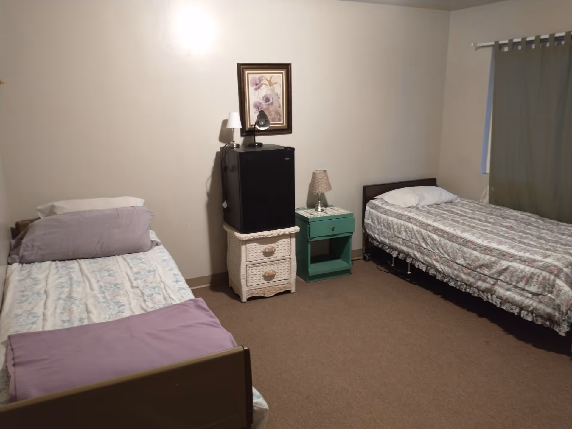 A simple bedroom with two single beds, each with floral patterned bedding and pillows. Between the beds are two small nightstands, one white wicker with a small black refrigerator on top, and the other green with a lamp. A framed floral picture hangs on the wall above the refrigerator. A window with green curtains is on the right wall.