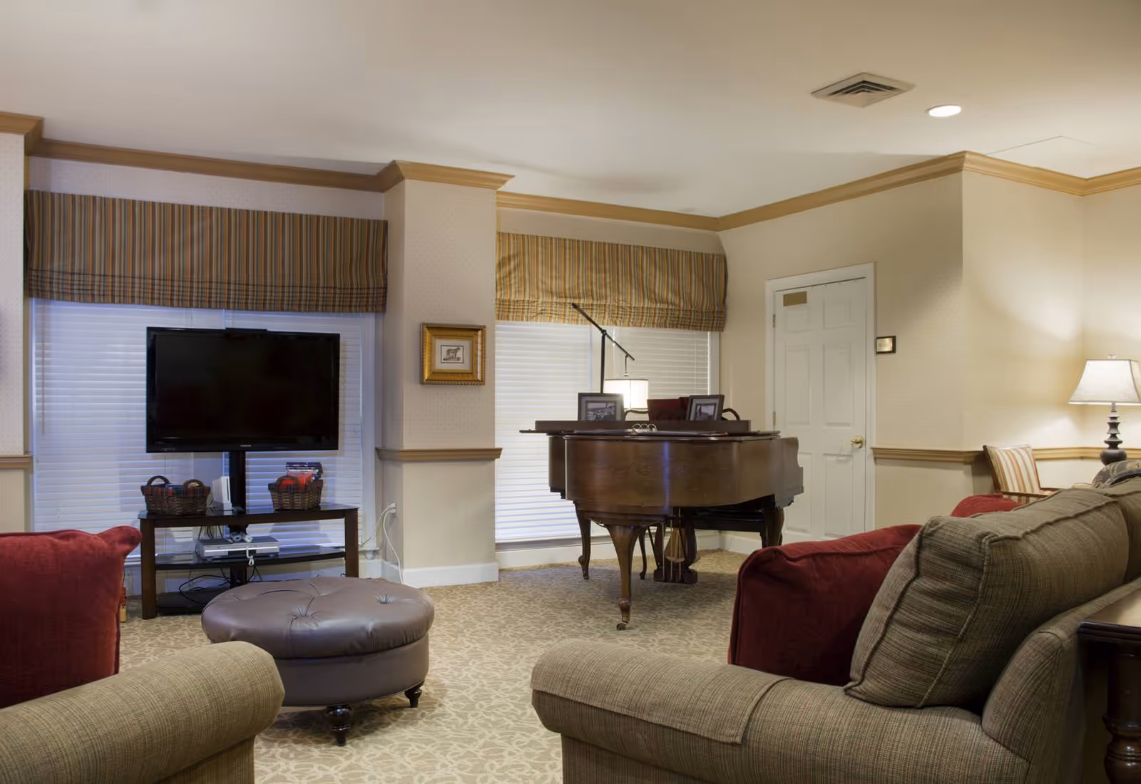 A cozy living room with beige walls and carpet, featuring a grand piano near two windows with striped valances. There is a flat-screen TV on a stand with baskets underneath, a round tufted ottoman, and several cushioned sofas and chairs with red and beige upholstery. A table lamp is visible on the right side of the room.