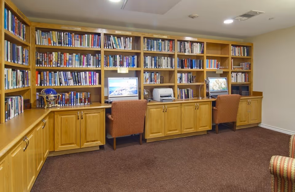 A cozy library or study area with wooden bookshelves filled with books, two computer workstations with desktop computers and chairs, a printer, and a globe on the left side. The room has carpeted flooring and neutral-colored walls.