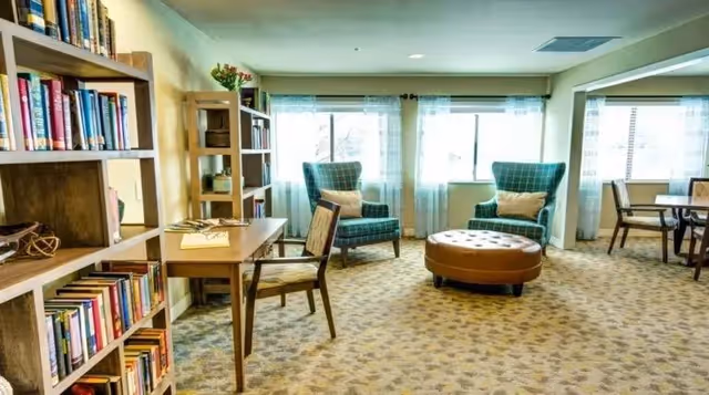 A bright and cozy common area with large windows letting in natural light. The room features two green plaid armchairs with beige pillows, a round brown ottoman, a wooden table with a chair, and bookshelves filled with books and decorative items. The carpet has a subtle patterned design.