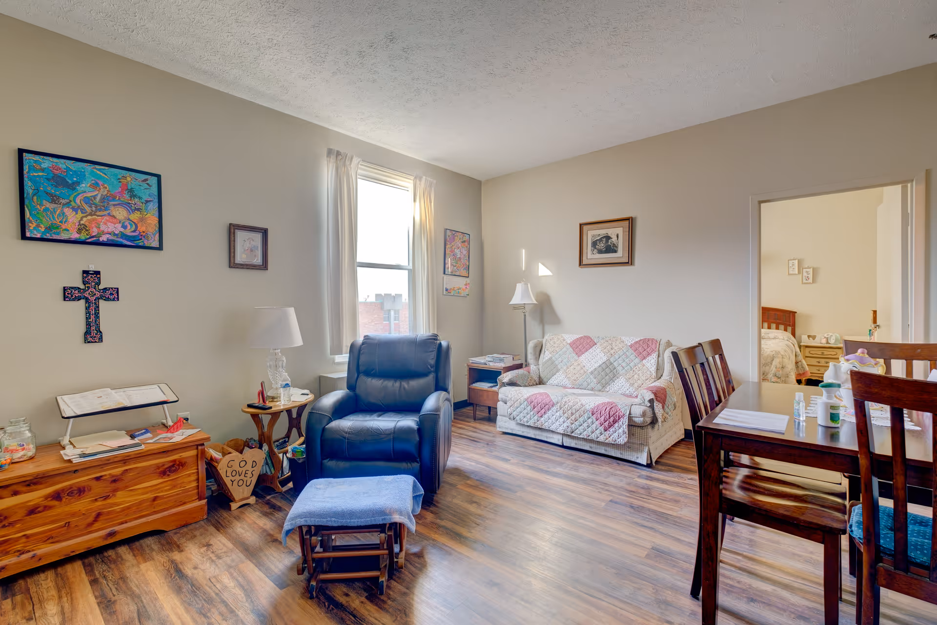 A cozy living room with a blue recliner chair and matching footrest, a wooden chest with a lamp and books, a small side table, a couch covered with a patchwork quilt, and a wooden dining table with chairs. The walls are decorated with colorful artwork and a cross. A doorway leads to a bedroom with a bed and nightstand visible.