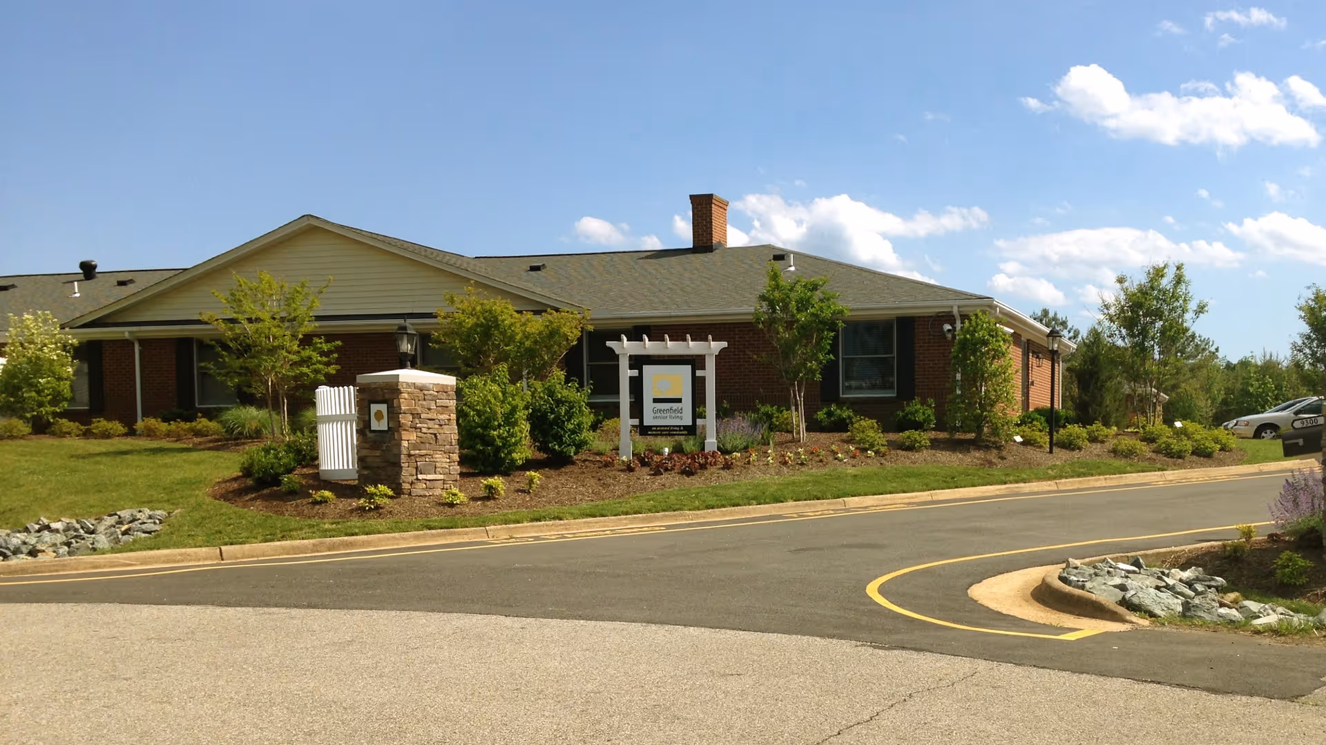 Exterior view of a single-story brick building with a green roof, surrounded by landscaped greenery and small trees under a blue sky with scattered clouds. A paved driveway curves in front of the building, and a sign is visible near the entrance.