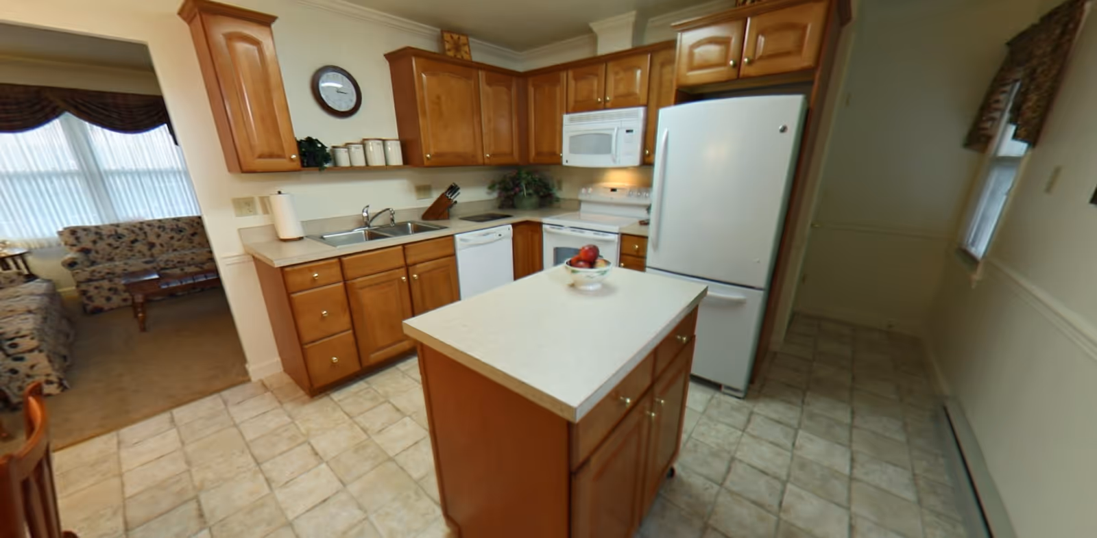 A kitchen with wooden cabinets, a white refrigerator, microwave, stove, dishwasher, and a small island with a bowl of fruit on top. The kitchen has tiled flooring and a double sink. Adjacent to the kitchen is a living room area with floral patterned sofas and a window with sheer curtains.