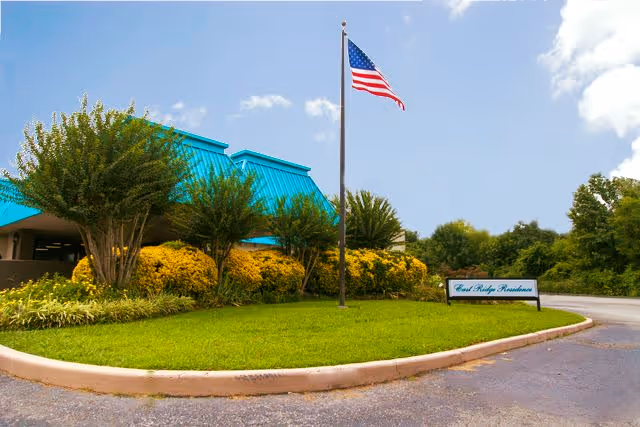 Exterior view of East Ridge Residence facility with a bright blue roof, manicured bushes, green lawn, and an American flag on a flagpole under a partly cloudy sky.