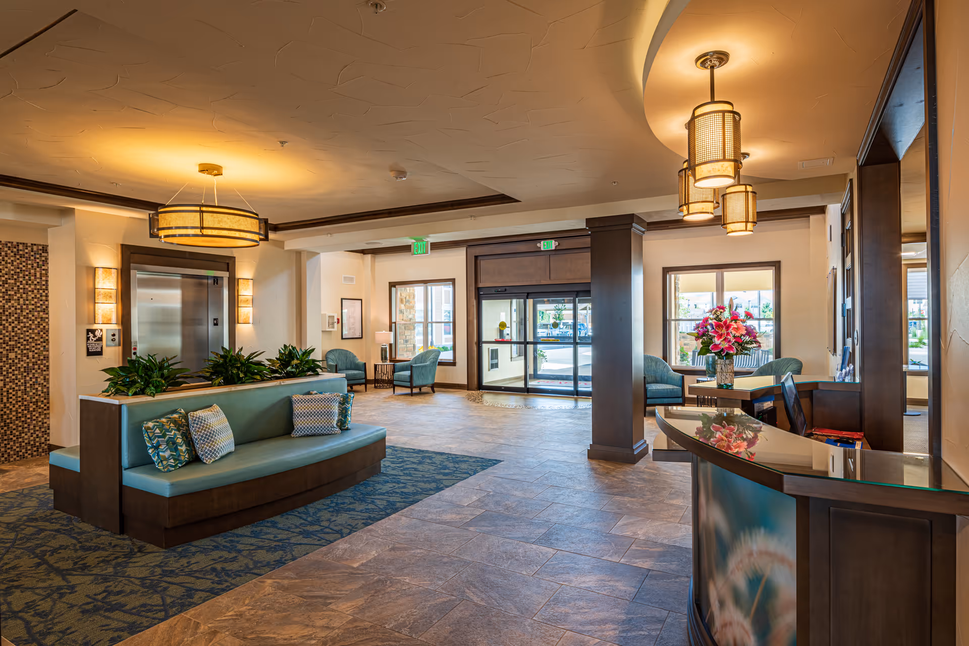 A spacious and well-lit lobby area with a seating bench adorned with decorative pillows, potted plants, and a reception desk with a flower arrangement. The lobby features large windows, an elevator, and comfortable chairs near the entrance with glass doors leading outside.