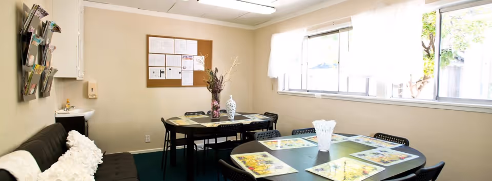 A small common room with two black tables, each surrounded by black chairs. The tables have placemats and decorative vases with flowers. There is a black couch with white pillows on the left side, a small sink, and a bulletin board on the beige wall. Large windows on the right side let in natural light and show some greenery outside.