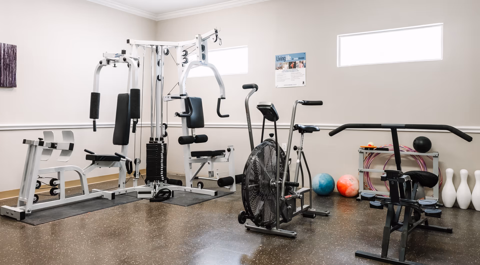 A small fitness room with various exercise equipment including a multi-station gym machine, an exercise bike, a rowing machine, and a stationary bike. There are also colorful exercise balls, hula hoops, and bowling pins arranged against the wall. The room has two rectangular windows near the ceiling and light-colored walls.
