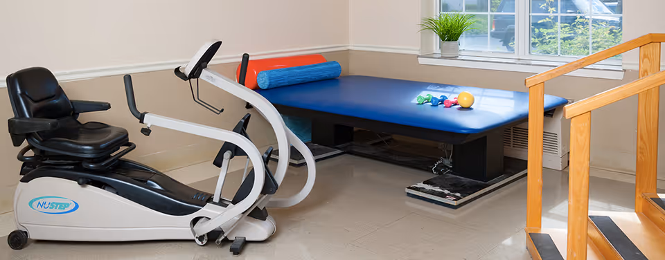 A small exercise room with a recumbent exercise bike, a padded therapy table with foam rollers and small dumbbells on it, and a wooden handrail leading up a few steps near a window.