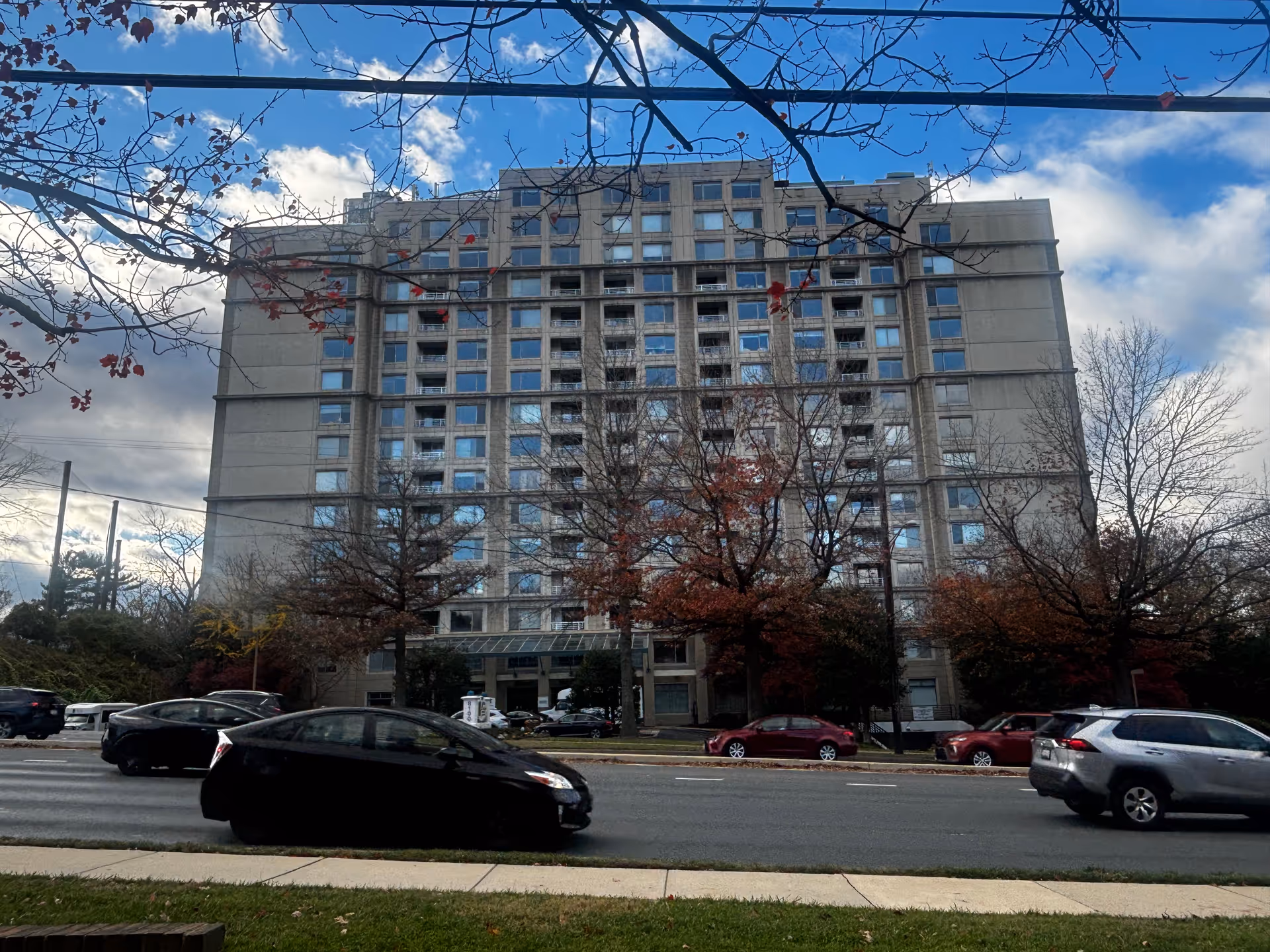 Exterior view of a multi-story residential building with numerous windows, trees with autumn leaves in front, and several cars driving on the road in the foreground under a partly cloudy sky.
