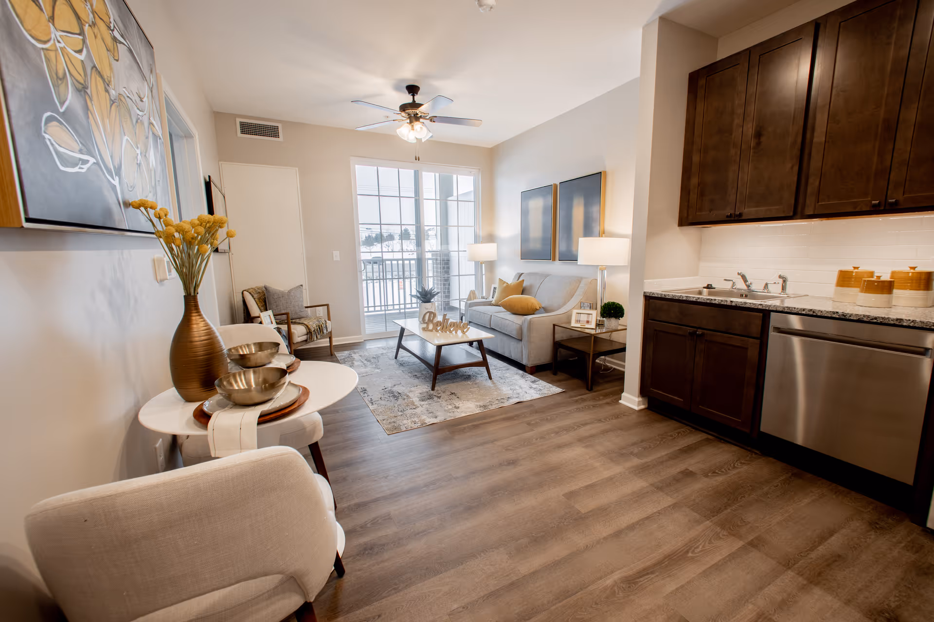 A modern senior living apartment interior featuring a small living room with a gray sofa, two framed artworks on the wall, a coffee table with a decorative 'Believe' sign, and two side tables with lamps. To the right is a kitchen area with dark wooden cabinets, a granite countertop, a stainless steel dishwasher, and a sink. The room has wood flooring, a ceiling fan with lights, and large windows letting in natural light. A small round table with a vase of yellow flowers and decorative bowls is in the foreground.