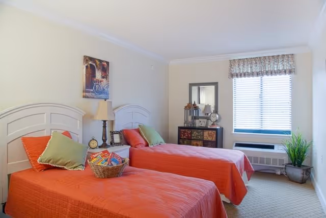 A bright bedroom with two twin beds featuring orange bedspreads and green pillows. Between the beds is a nightstand with a lamp, an alarm clock, and a basket with magazines. On the far wall, there is a window with blinds and a floral valance, a small dresser with decorative items, and a potted plant on the floor.