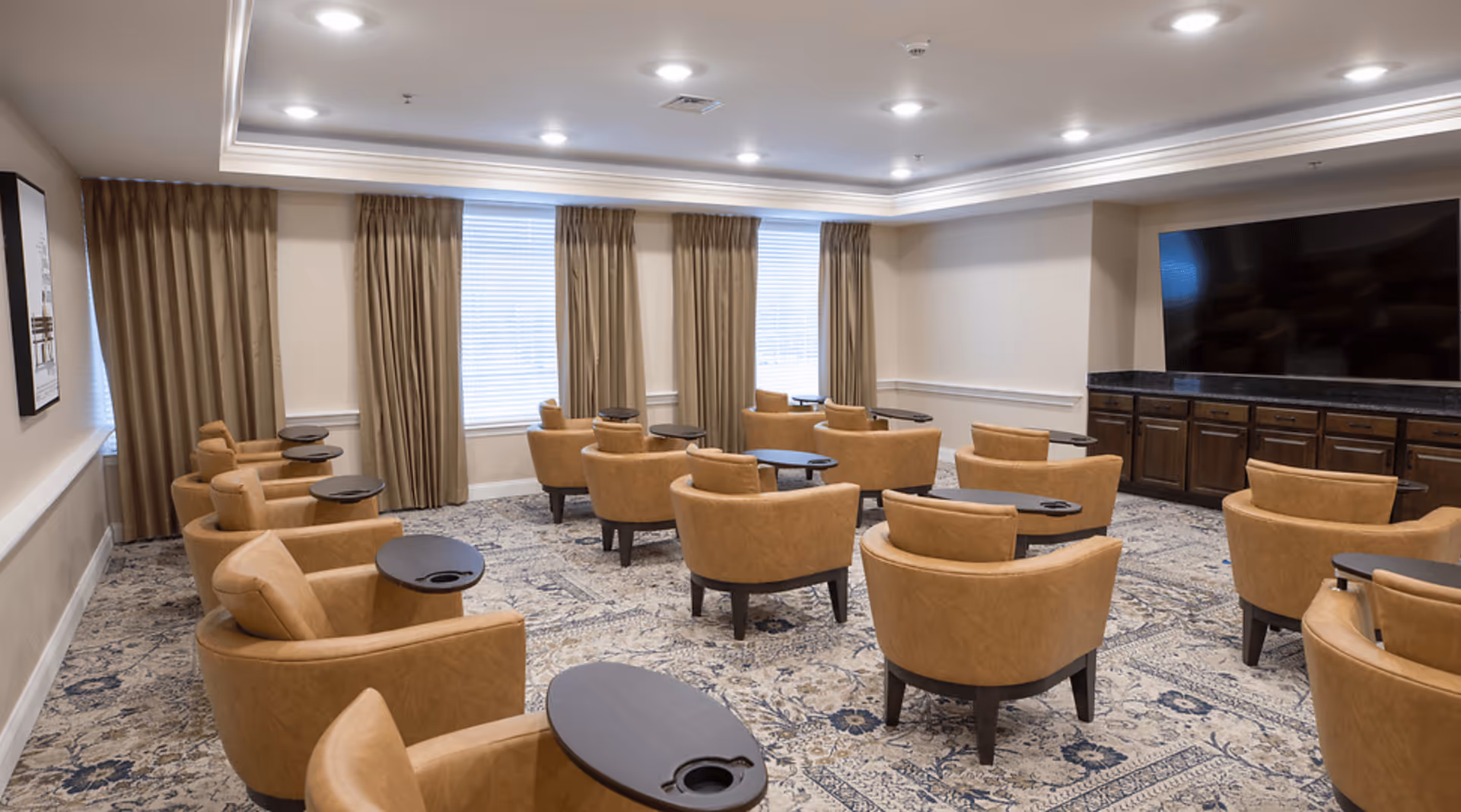 A spacious room with multiple tan leather armchairs arranged in rows, each with a small attached black table. The room has large windows with beige curtains, a patterned carpet, and a large flat-screen TV mounted on a dark wooden cabinet against one wall. The ceiling has recessed lighting and crown molding.