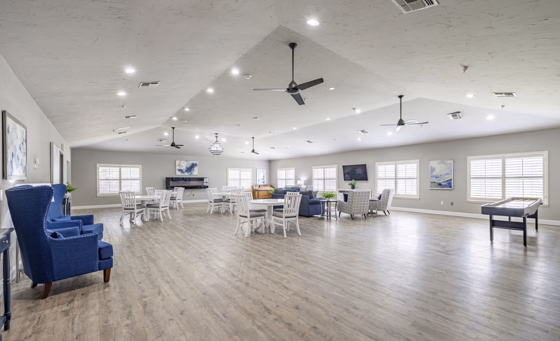A spacious, well-lit common area in a senior living facility with light wood flooring, white walls, and multiple windows with white shutters. The room features several round tables with white chairs, blue upholstered armchairs, a seating area with patterned chairs and a sofa, ceiling fans, and a shuffleboard table. Artwork and plants decorate the space.