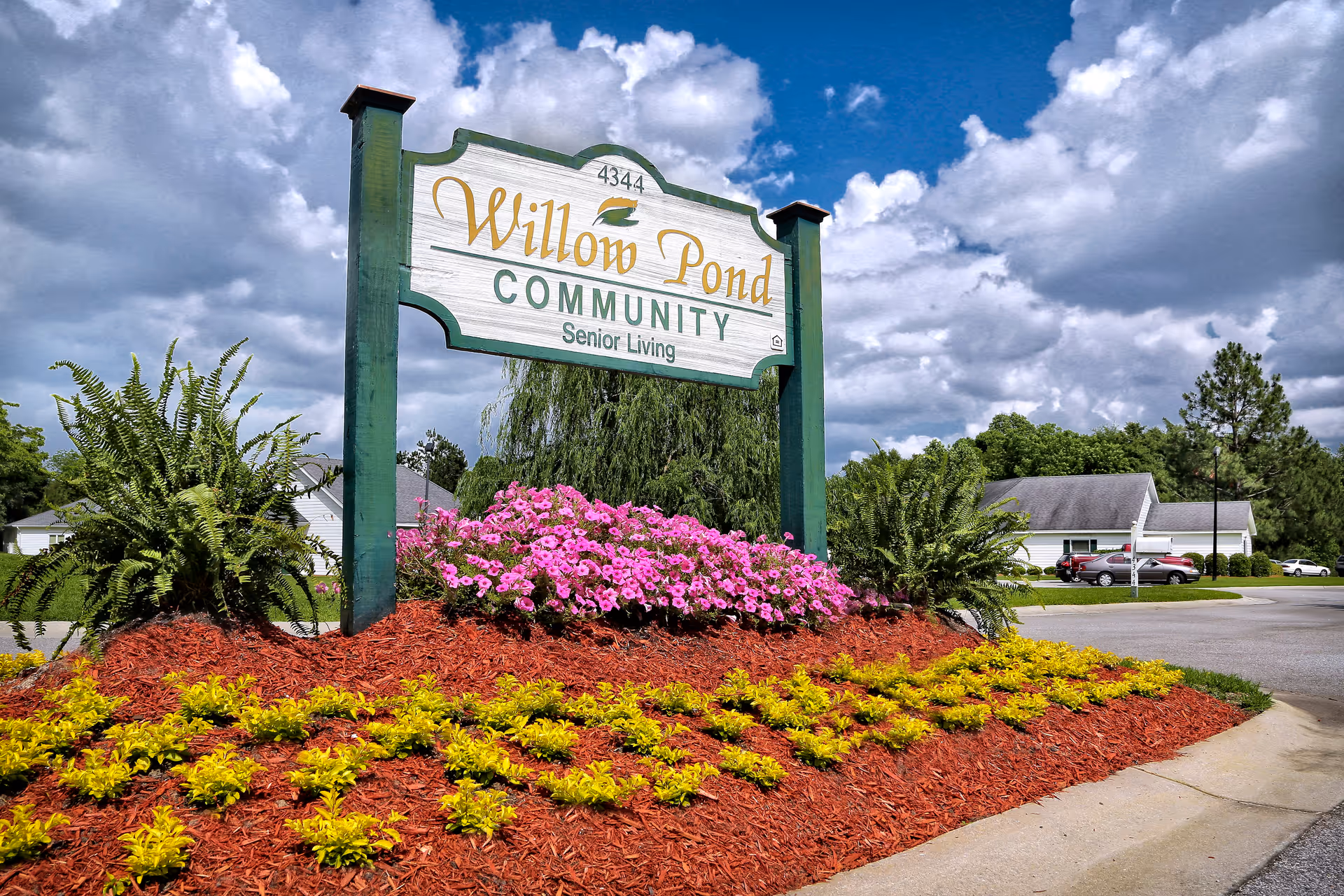 Outdoor view of a landscaped entrance sign for Willow Pond Community Senior Living, surrounded by colorful flowers and greenery under a partly cloudy sky.