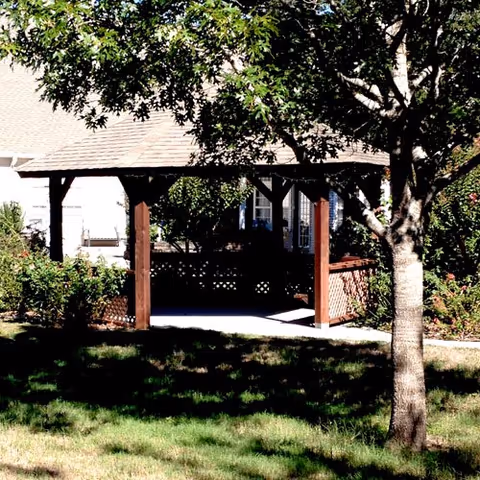 A wooden gazebo with a shingled roof situated in a garden area with green grass, bushes, and a tree in the foreground. The gazebo has lattice panels and is surrounded by greenery.