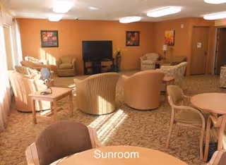 A sunroom with multiple beige and tan upholstered chairs and sofas arranged around a large flat-screen TV mounted on a stand. There are small tables with lamps and floral decorations, and the room is warmly lit with natural light coming through large windows on the left side. The walls are painted a warm orange color with framed artwork hanging on them. The carpet is patterned in neutral tones.