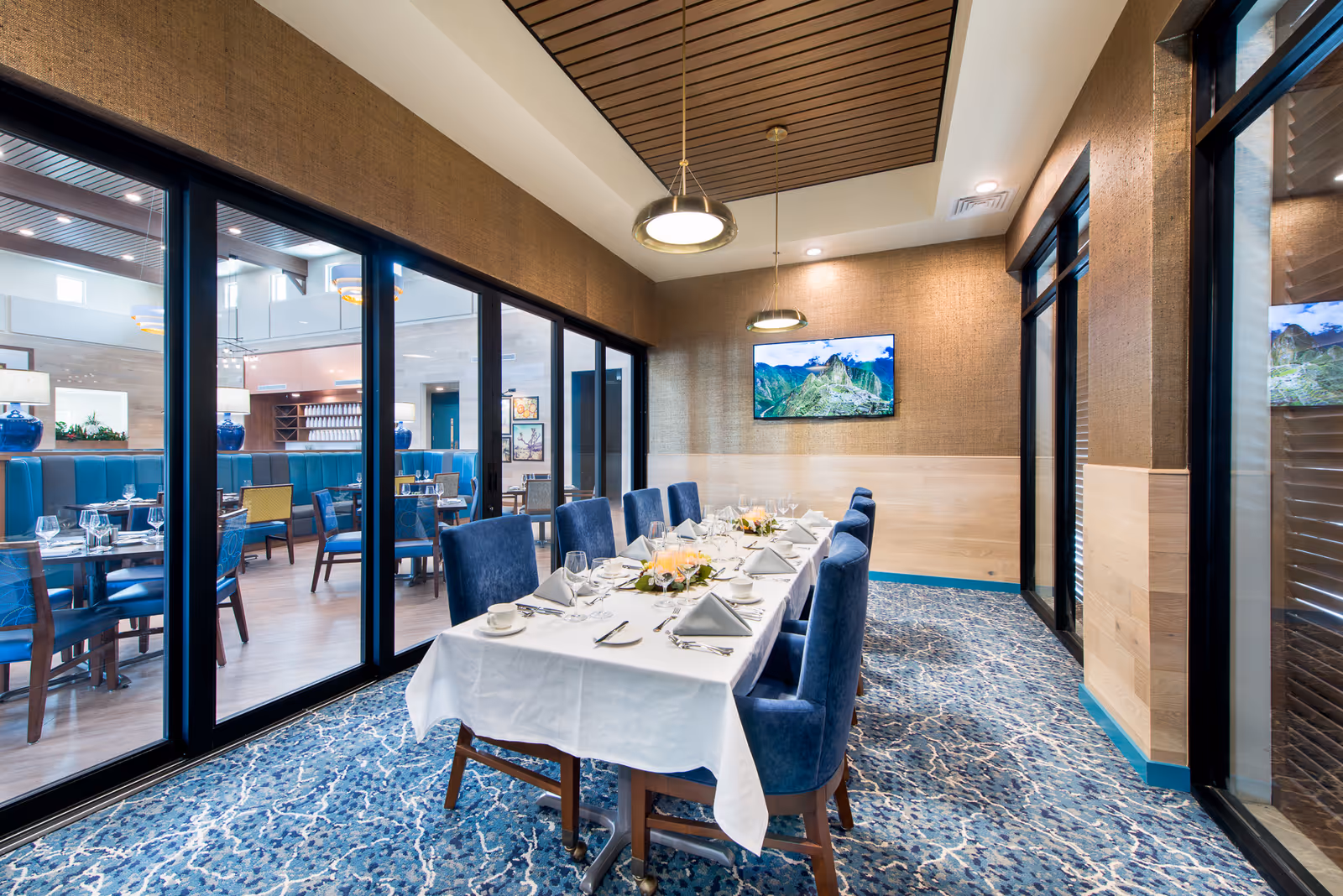 Private dining room with a long white-tableclothed table set for a meal, blue upholstered chairs, and a glass wall overlooking the main dining area.