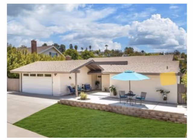 Single-story house with a beige exterior and a two-car garage, featuring a front patio with outdoor seating and a blue umbrella, surrounded by a green lawn and trees under a partly cloudy sky.