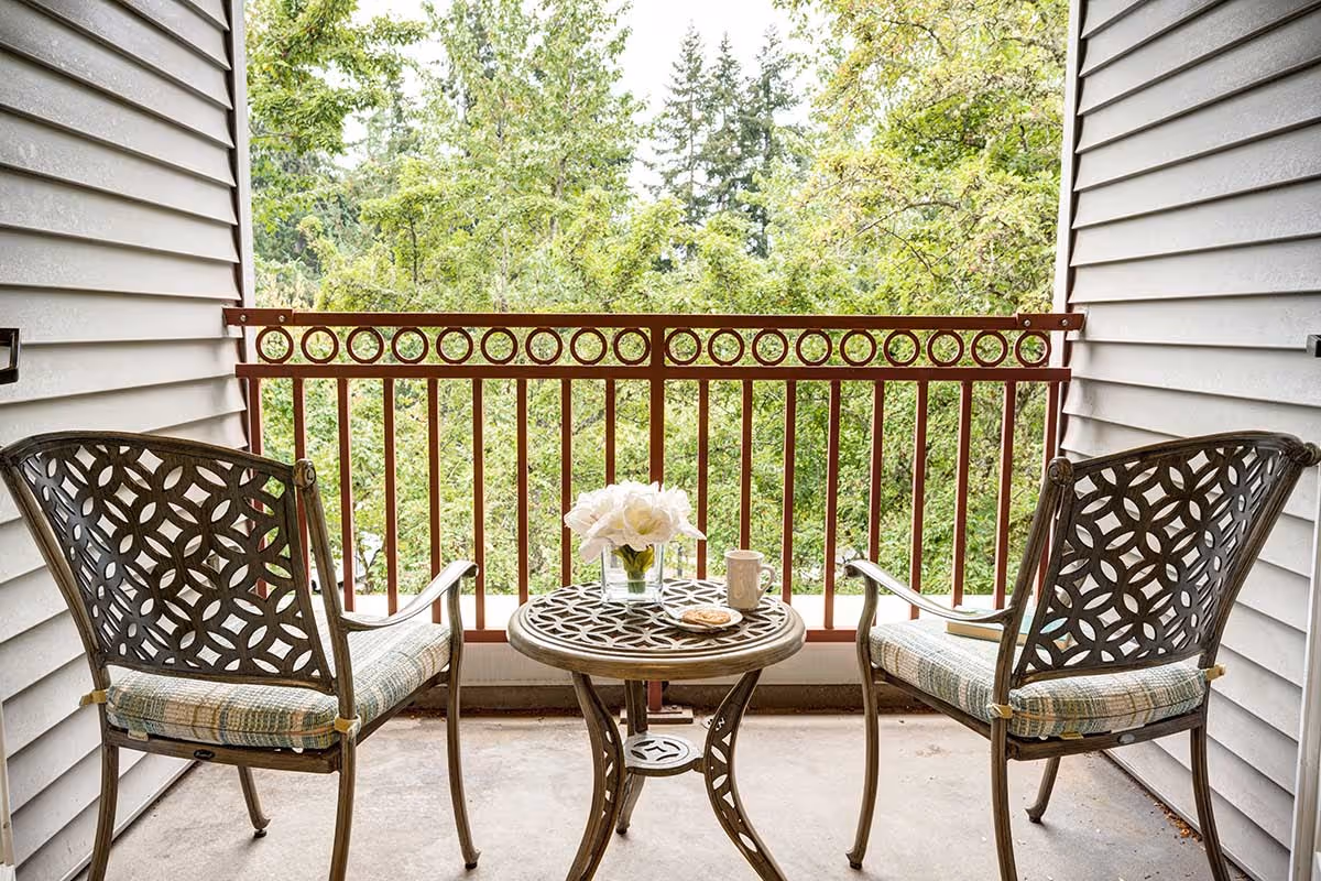 A small outdoor balcony area with two metal chairs featuring patterned cushions and a matching round table between them. On the table, there is a small vase with white flowers, a coffee mug, and a plate with a cookie. The balcony has a red metal railing and overlooks green trees and foliage.